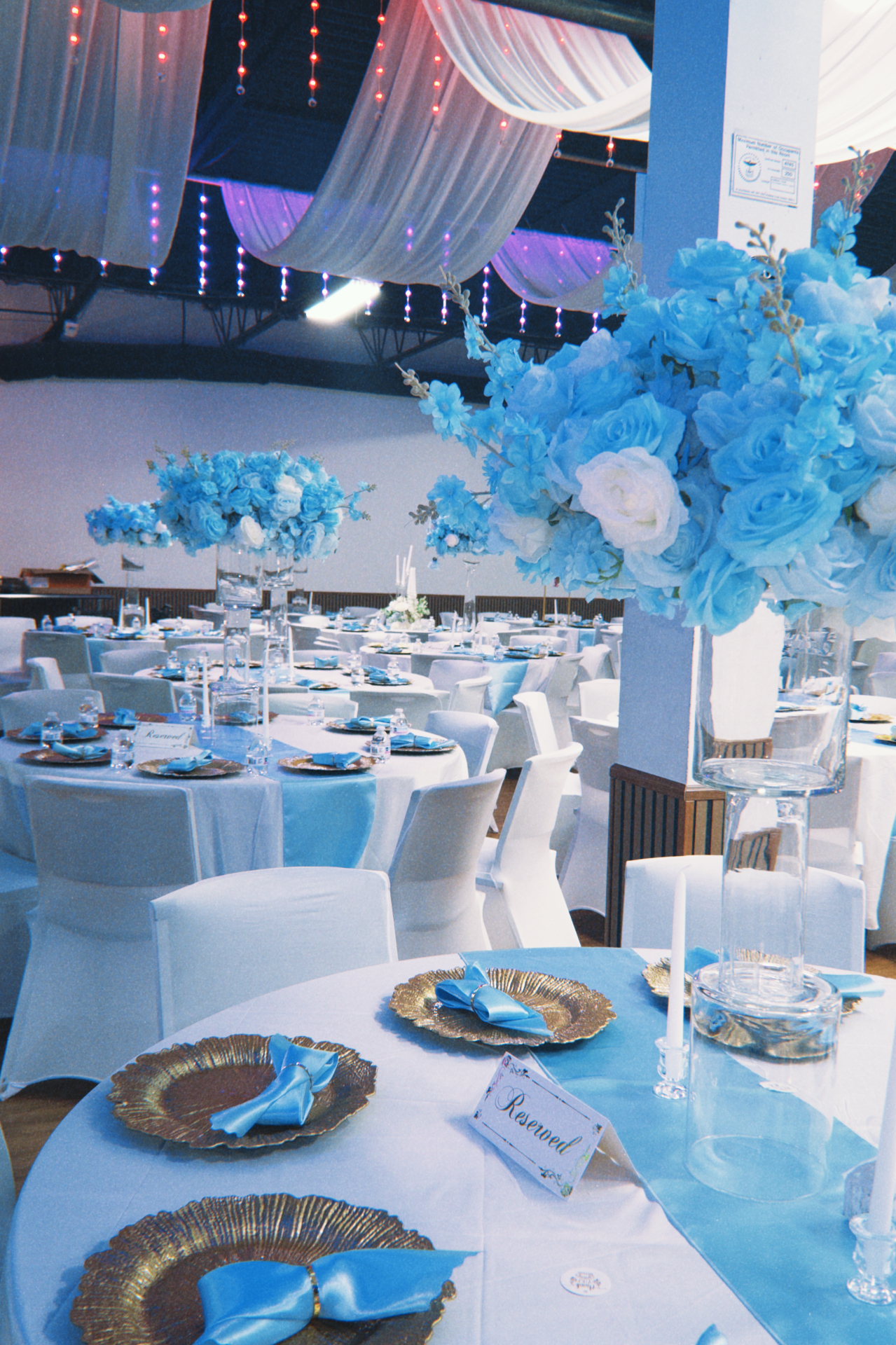 Wedding reception hall with rows of round tables, blue florals, draped ceiling, and ambient lighting.